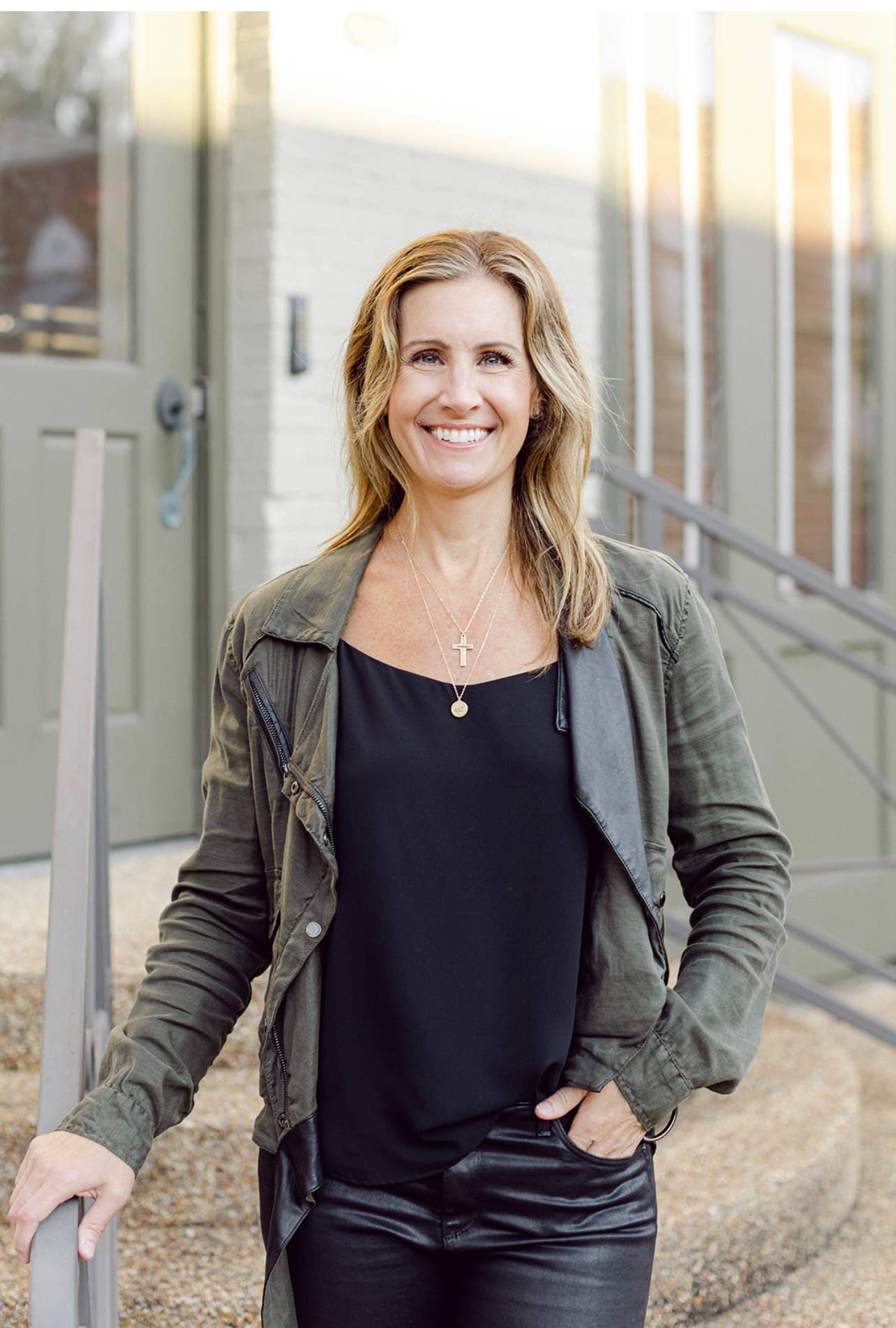 Woman standing outdoors wearing a green jacket and black top, smiling.