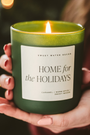 Hand holding a 'Home for the Holidays' candle with a blurred Christmas tree in the background
