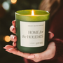 Hand holding a 'Home for the Holidays' candle with a blurred Christmas tree in the background
