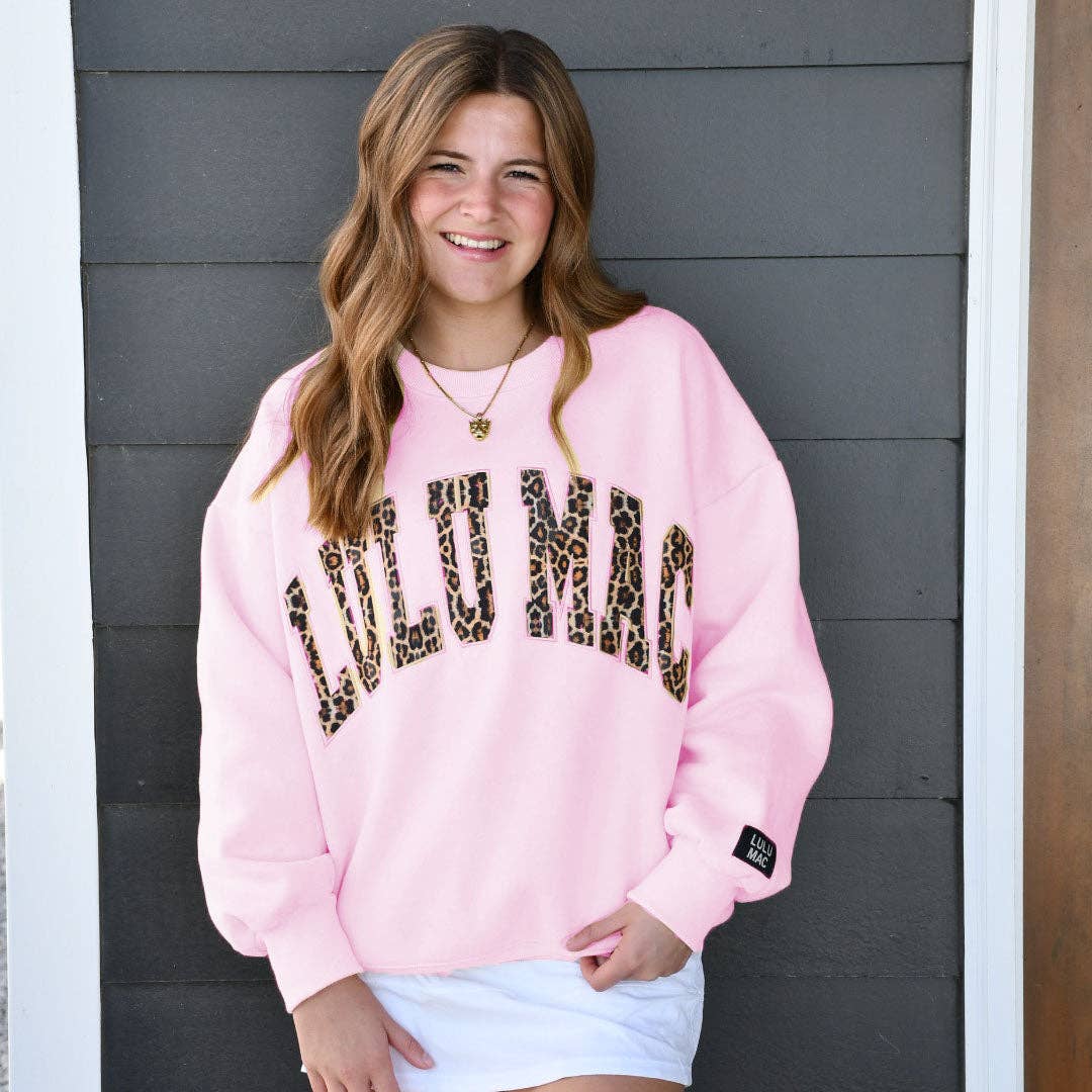 Person wearing a pink sweatshirt with leopard print text against a gray wall.