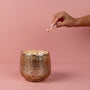 Hand lighting a candle in a textured gold container against a pink background