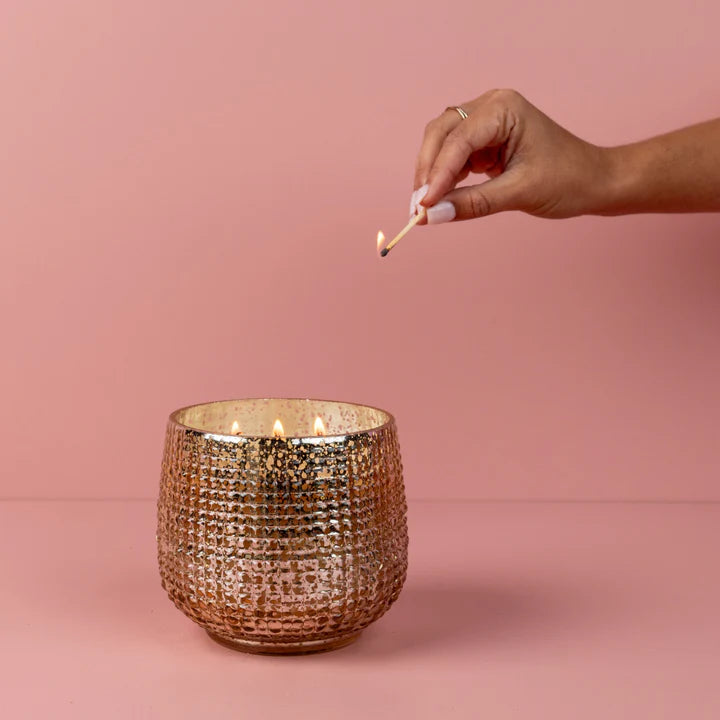 Hand lighting a candle in a textured gold container against a pink background