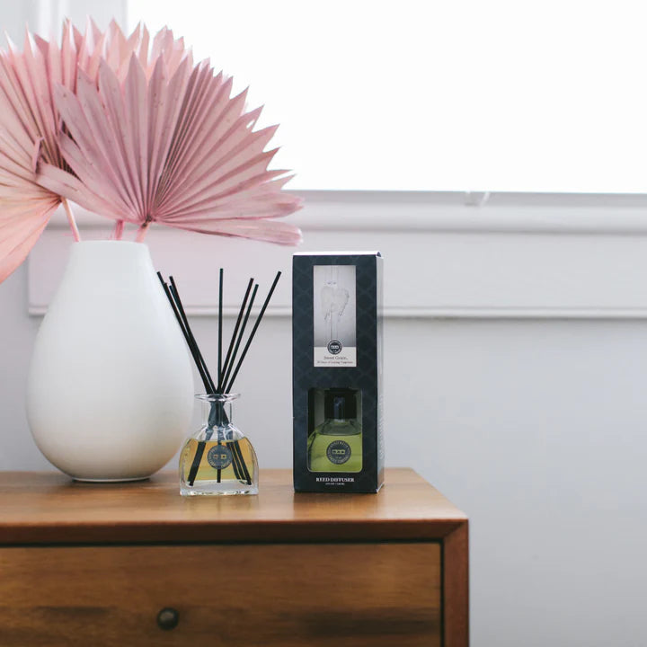 A diffuser with reeds and packaging on a wooden surface with a white wall in the background.