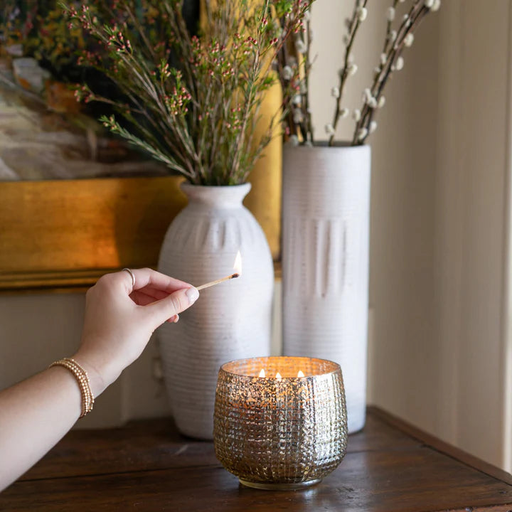 Person lighting a candle in a textured metal holder with decorative vases and plants in the background.