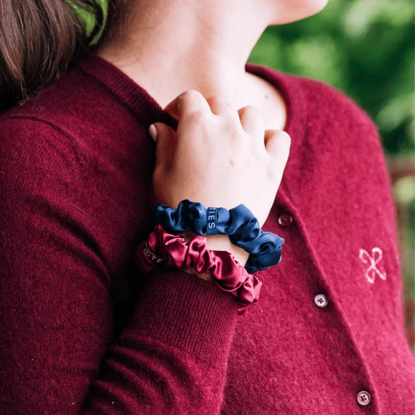 A close-up of a person wearing three satin scrunchie bracelets in navy blue, dark pink, and green, styled with a burgundy knit cardigan.