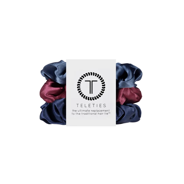 A set of three Teleties hair scrunchies in navy blue, dusty blue, and burgundy, wrapped together with a white paper label featuring the Teleties logo.