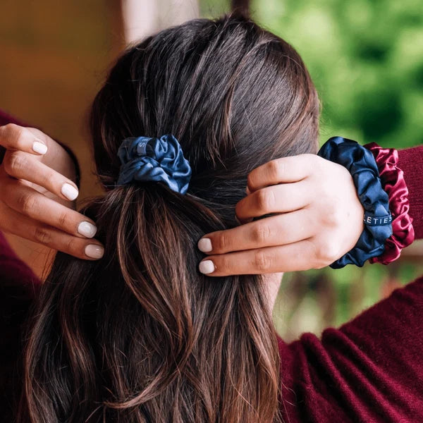 A close-up lifestyle shot of a person from behind, wearing three satin scrunchies in navy blue, dark blue, and burgundy on their brown hair. The brand name 'ETIE' is partially visible on the burgundy scrunchie.
