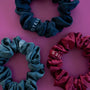Three satin fabric hair scrunchies in dark teal, dusty blue, and burgundy red, arranged on a pink background. Each scrunchie has a white embroidered brand tag reading 'TELLIES'.