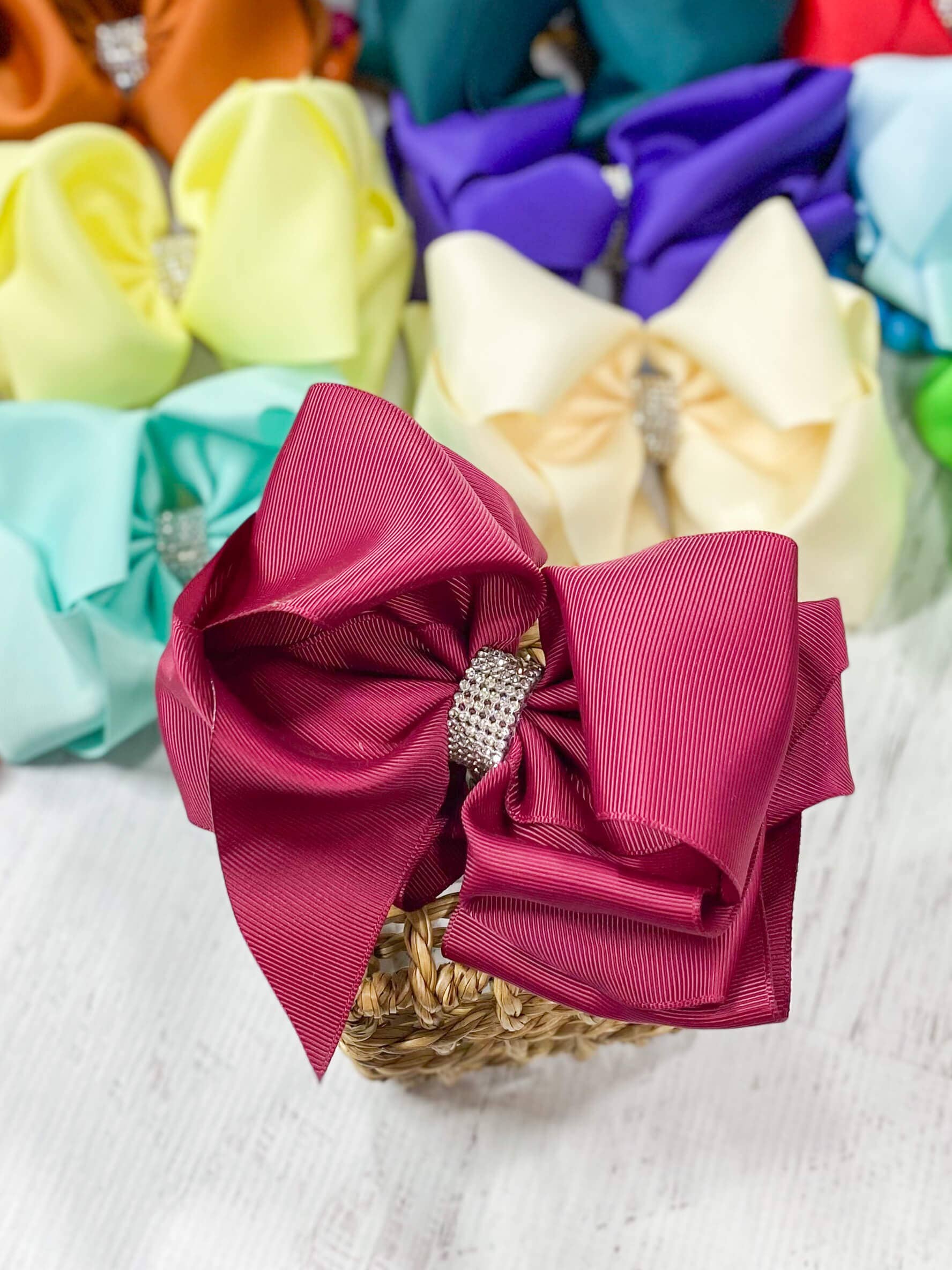 Large pink bow with a rhinestone center on a textured surface with other colorful bows in the background.