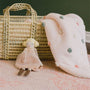 Small doll in a pink dress next to a textured blanket and wicker basket on a pink fabric background.