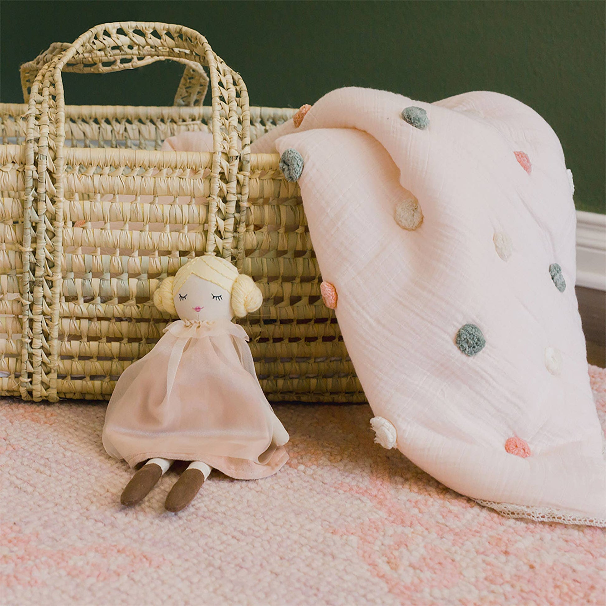 Small doll in a pink dress next to a textured blanket and wicker basket on a pink fabric background.