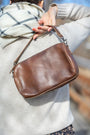Brown leather handbag held by a person wearing a light-colored sweater and plaid scarf.
