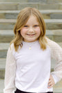 Young girl with blonde hair wearing a white shirt with sheer sleeves against a stone wall background