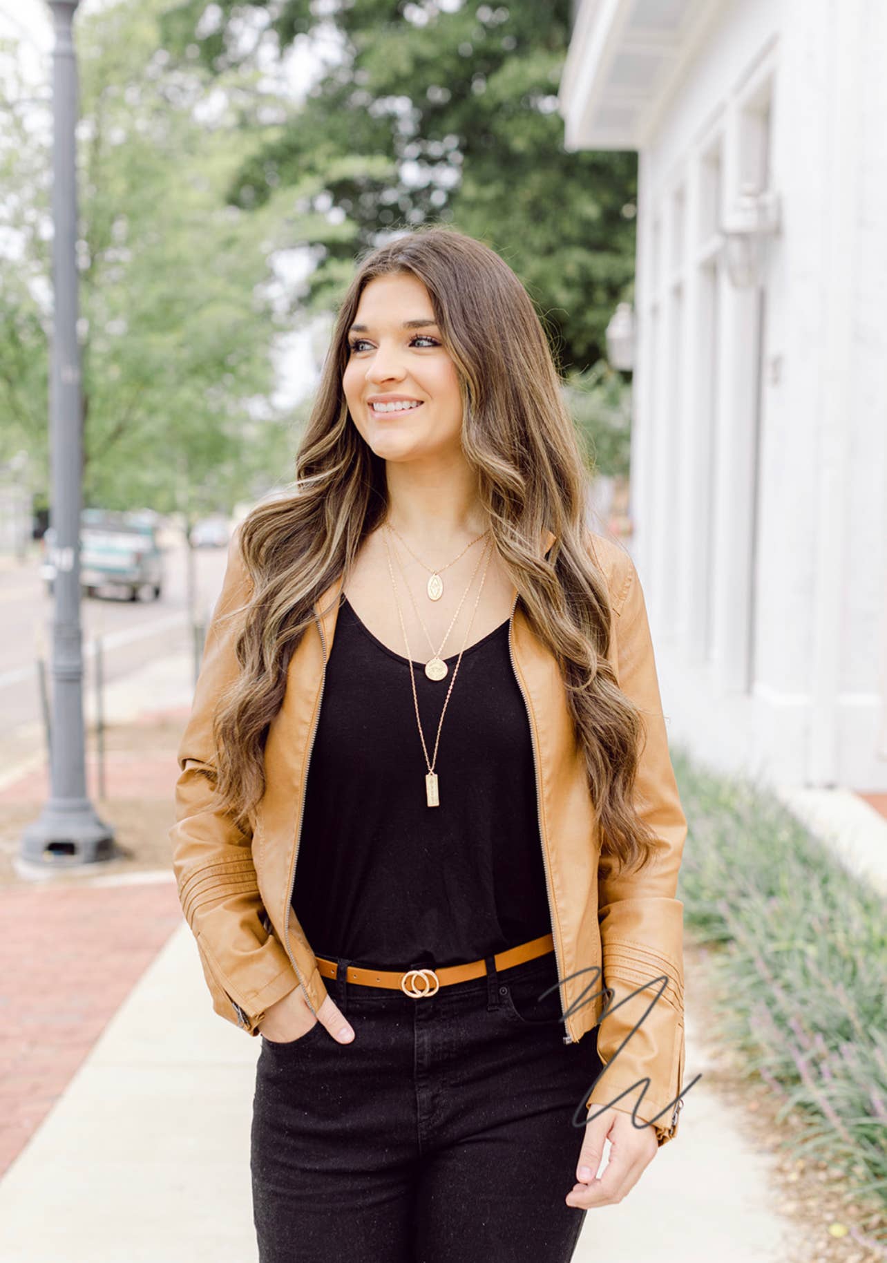 Woman wearing a tan jacket and black outfit with a necklace, standing outdoors.