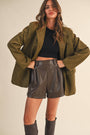 Woman wearing an olive green jacket, black top, and leather shorts on a beige background