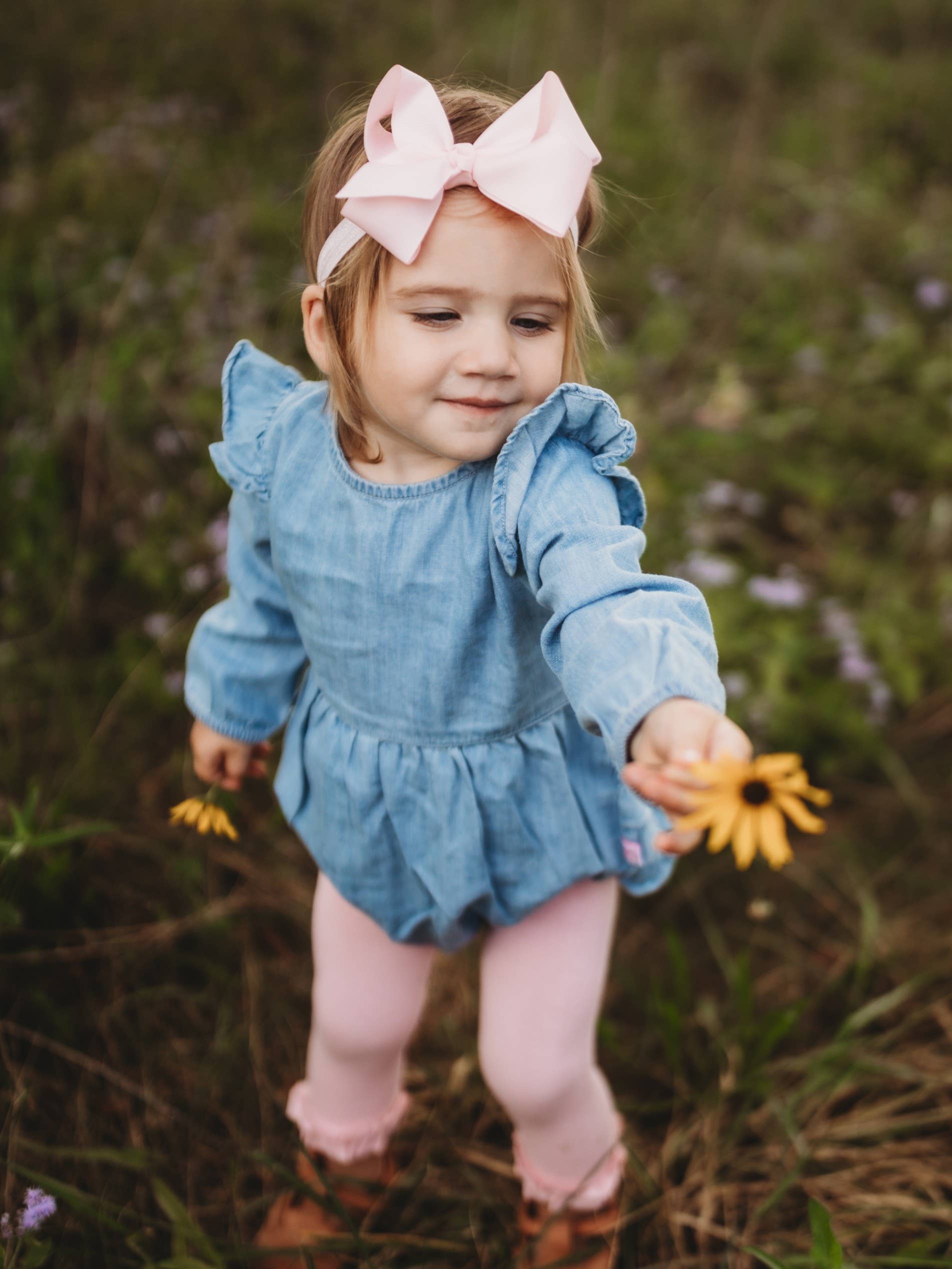 Child in a blue dress with a large pink bow standing in a field of flowers.