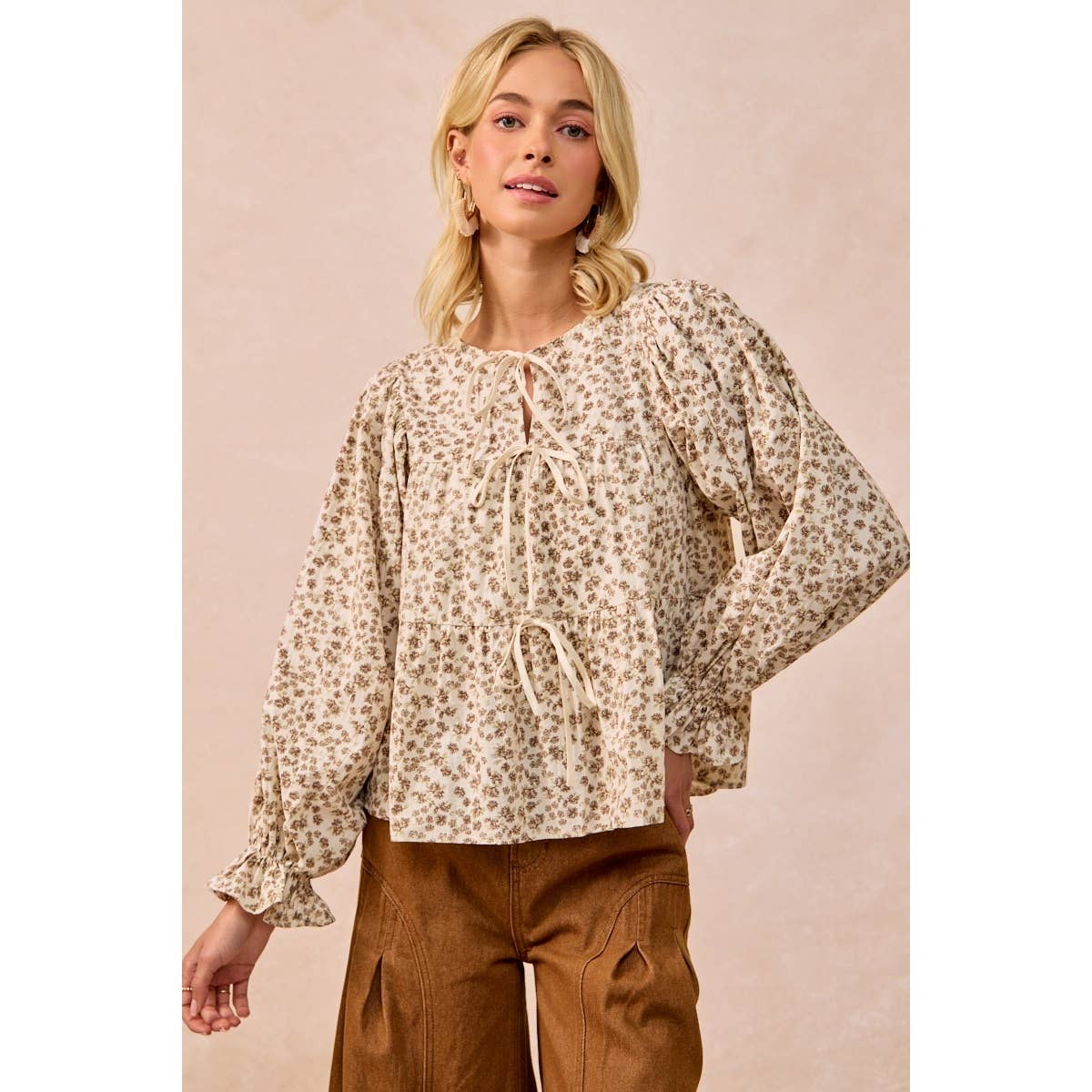 A woman modeling a beige long-sleeve blouse with a small brown floral print, featuring a tie-neck detail and puff sleeves, paired with brown pants against a solid beige background.
