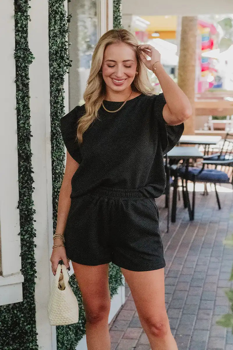 Woman wearing a black romper standing outdoors near a building with greenery.