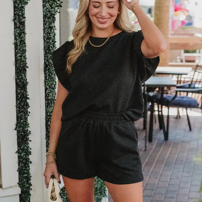 Woman wearing a black romper standing outdoors near a building with greenery.