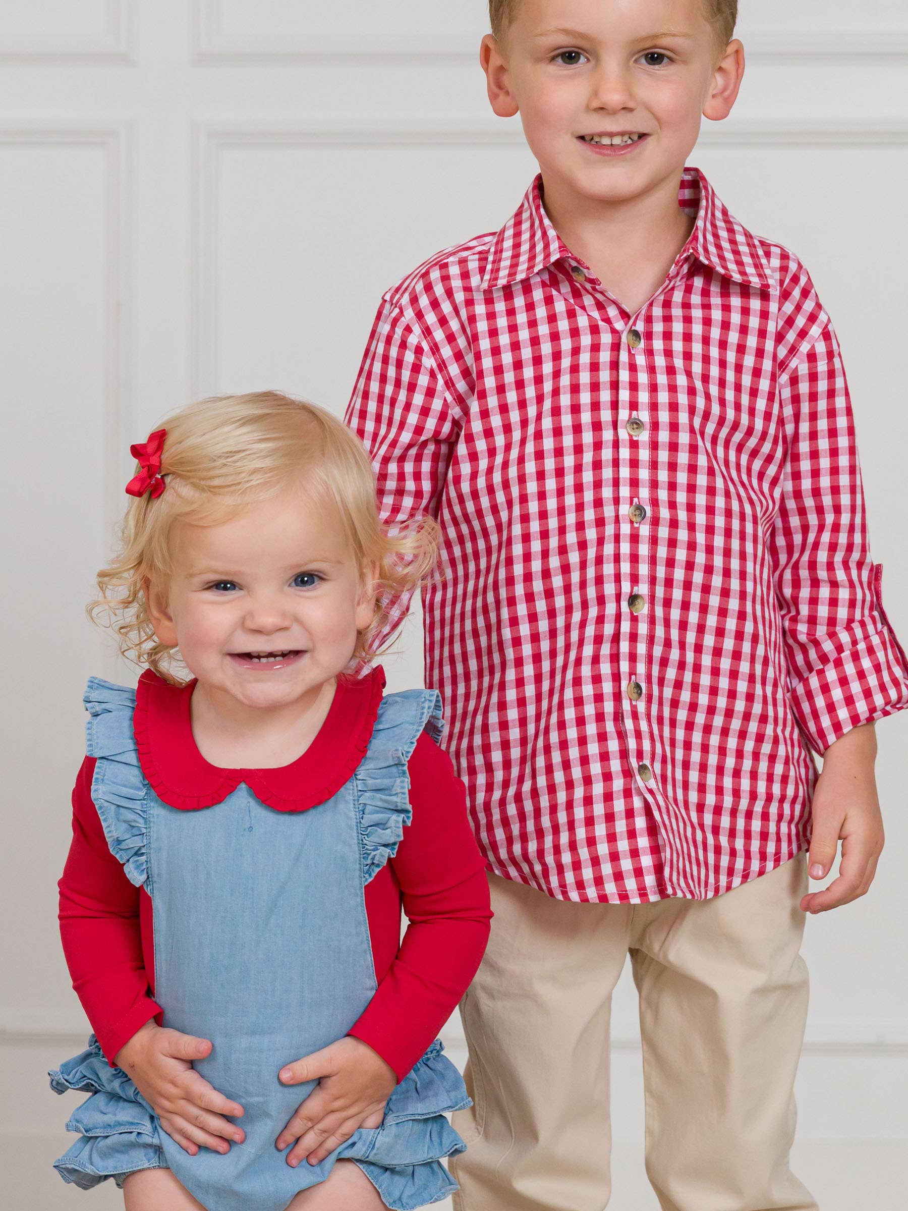 Two children, a girl in a blue dress with red collar and a boy in a red and white checkered shirt and beige pants, standing against a white background.