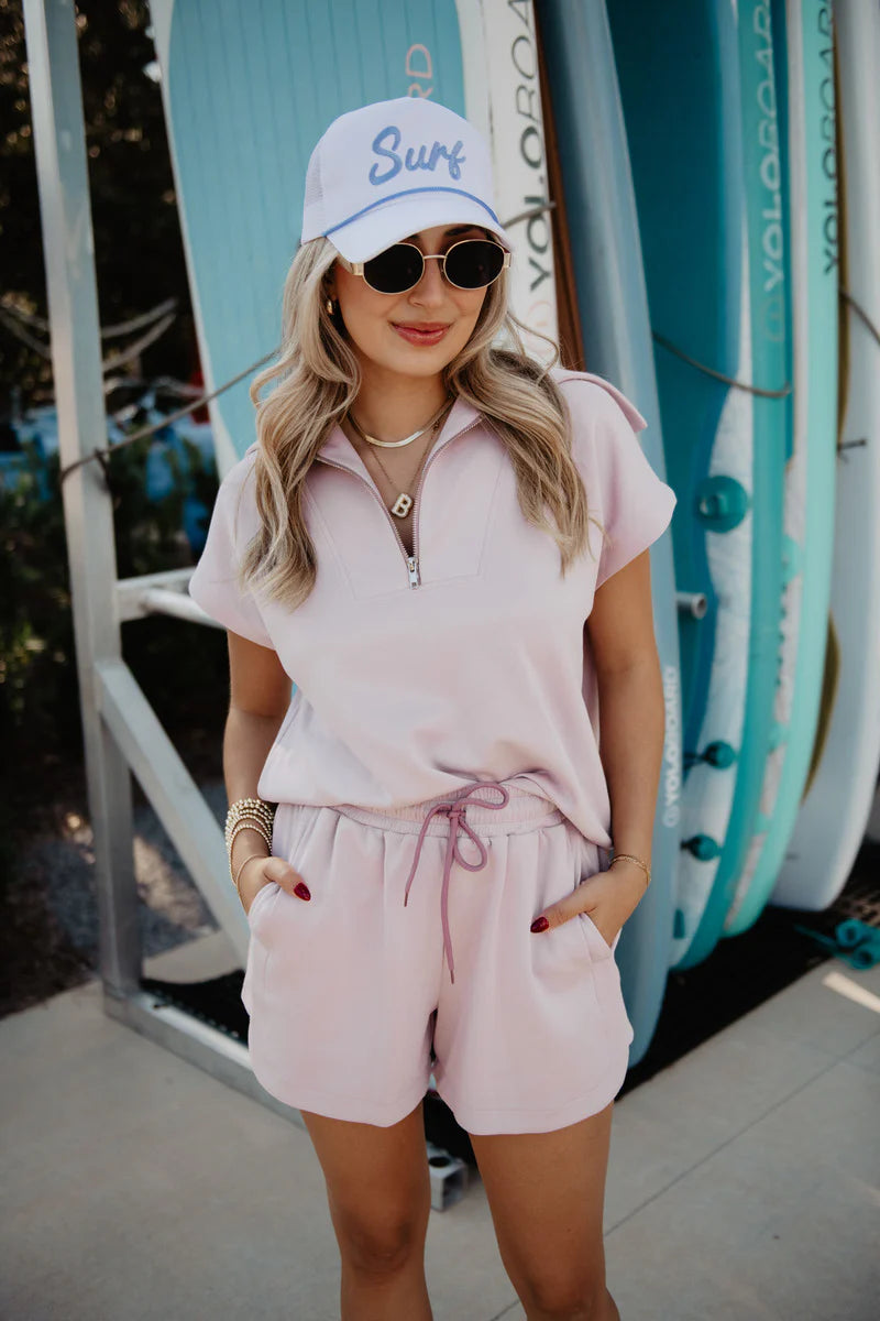 Woman wearing a pink outfit with sunglasses and a cap, standing in front of surfboards.