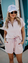 Woman wearing a pink outfit with sunglasses and a cap, standing in front of surfboards.