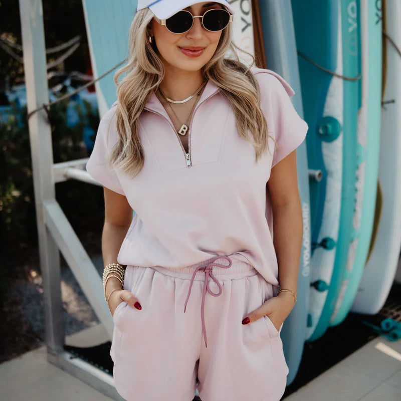 Woman wearing a pink outfit with sunglasses and a cap, standing in front of surfboards.