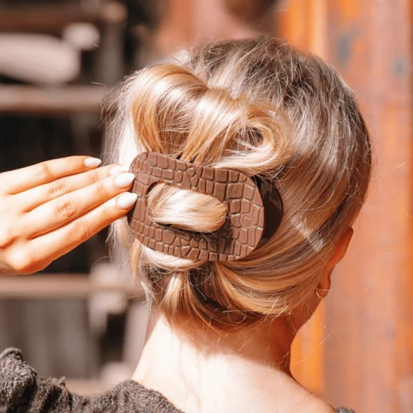 A close-up lifestyle shot of a woman from behind, showing her blonde hair styled in a low bun secured with a large, textured brown claw hair clip. A hand is adjusting the clip, and the background is a softly blurred warm-toned indoor setting.