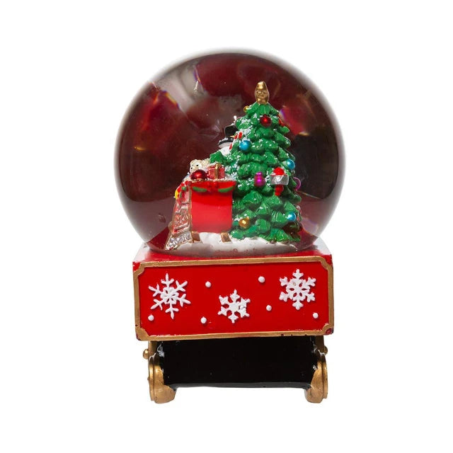 A Christmas-themed snow globe figurine featuring a green decorated tree, a red gift sack, and a red base with white snowflakes, set against a plain white background.