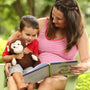 Woman reading a book to a child holding a monkey plush toy outdoors.