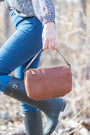 Person holding a brown leather bag outdoors with blurred natural background