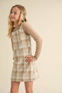 Young girl wearing a plaid dress over a long-sleeve shirt against a beige background