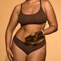 Person wearing a brown bikini holding a chocolate donut against a beige background