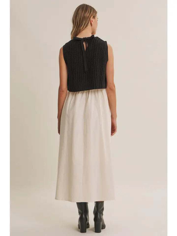 A woman shown from the back wearing a sleeveless black ribbed knit top with a tie detail at the neckline and a long cream-colored skirt, paired with black boots, against a plain light beige studio background.