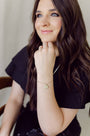 Woman with long brown hair wearing a black top and gold bracelet, sitting indoors.