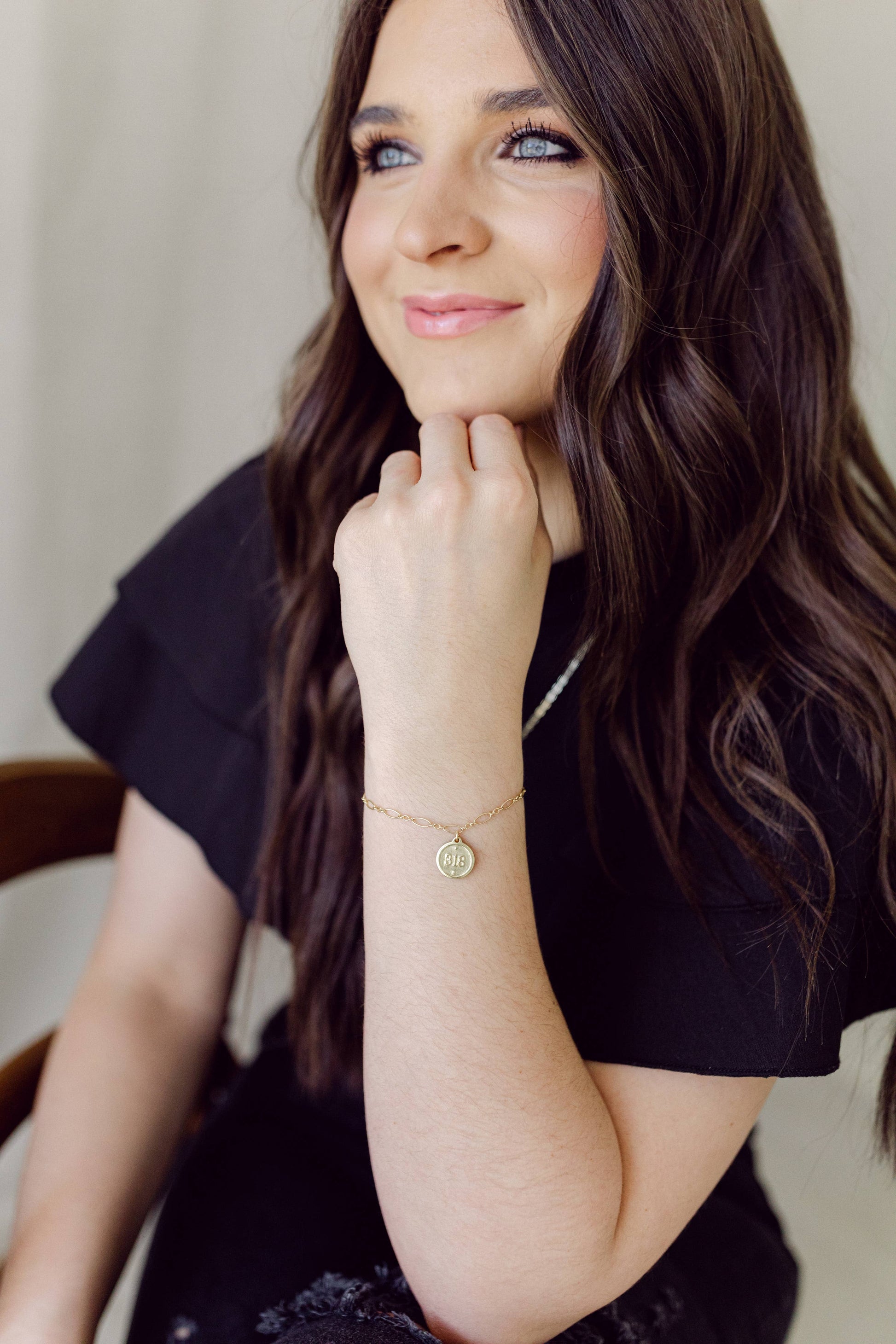 Woman with long brown hair wearing a black top and gold bracelet, sitting indoors.