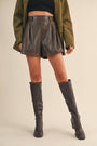 Person wearing a green coat, brown leather shorts, and knee-high boots on a beige background