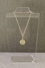 A gold chain necklace with a round medallion pendant displayed on a clear acrylic stand against a textured gray background.