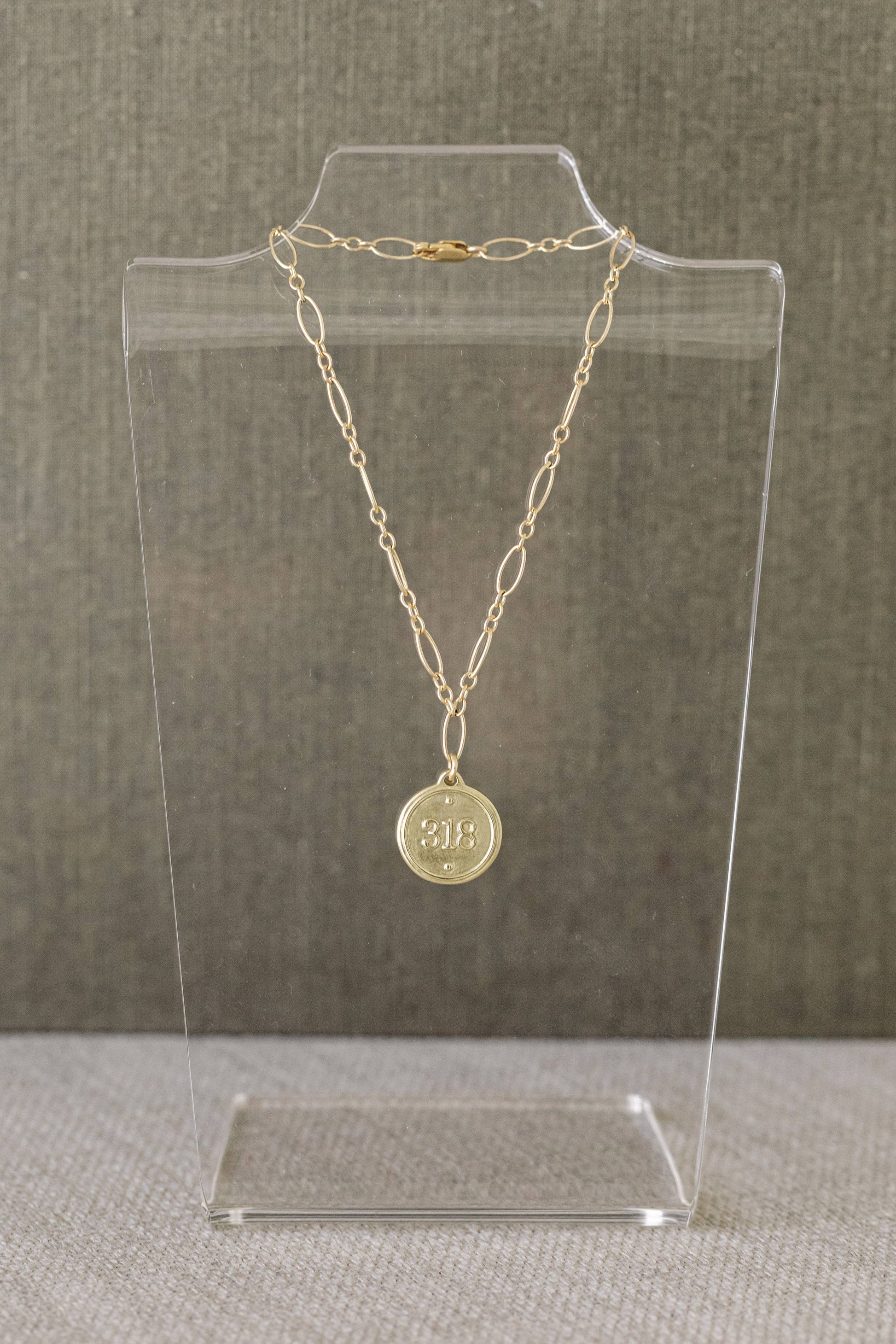 A gold chain necklace with a round medallion pendant displayed on a clear acrylic stand against a textured gray background.