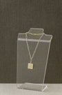 A delicate gold chain necklace with a square pendant engraved with '82$', displayed on a clear acrylic stand against a dark textured background.