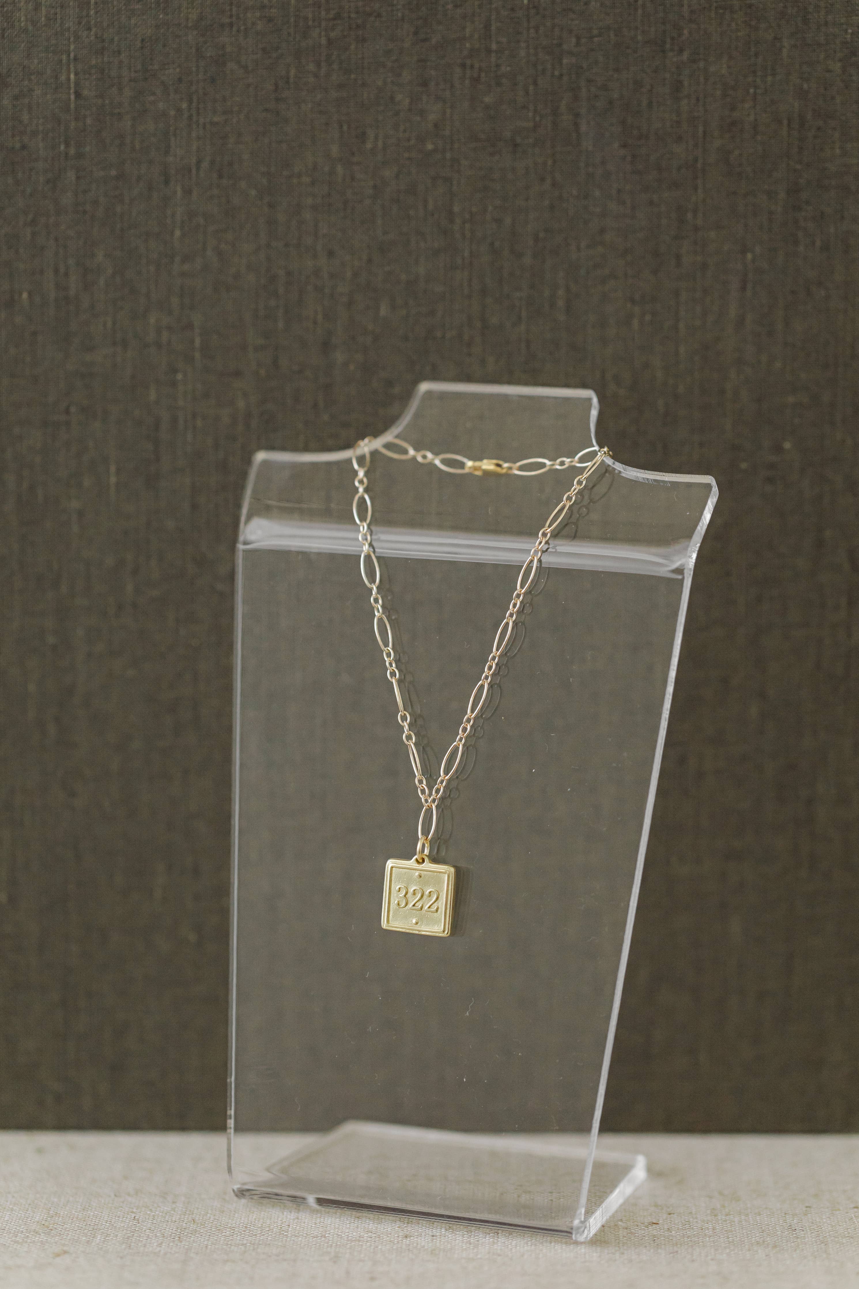 A delicate gold chain necklace with a square pendant engraved with '82$', displayed on a clear acrylic stand against a dark textured background.