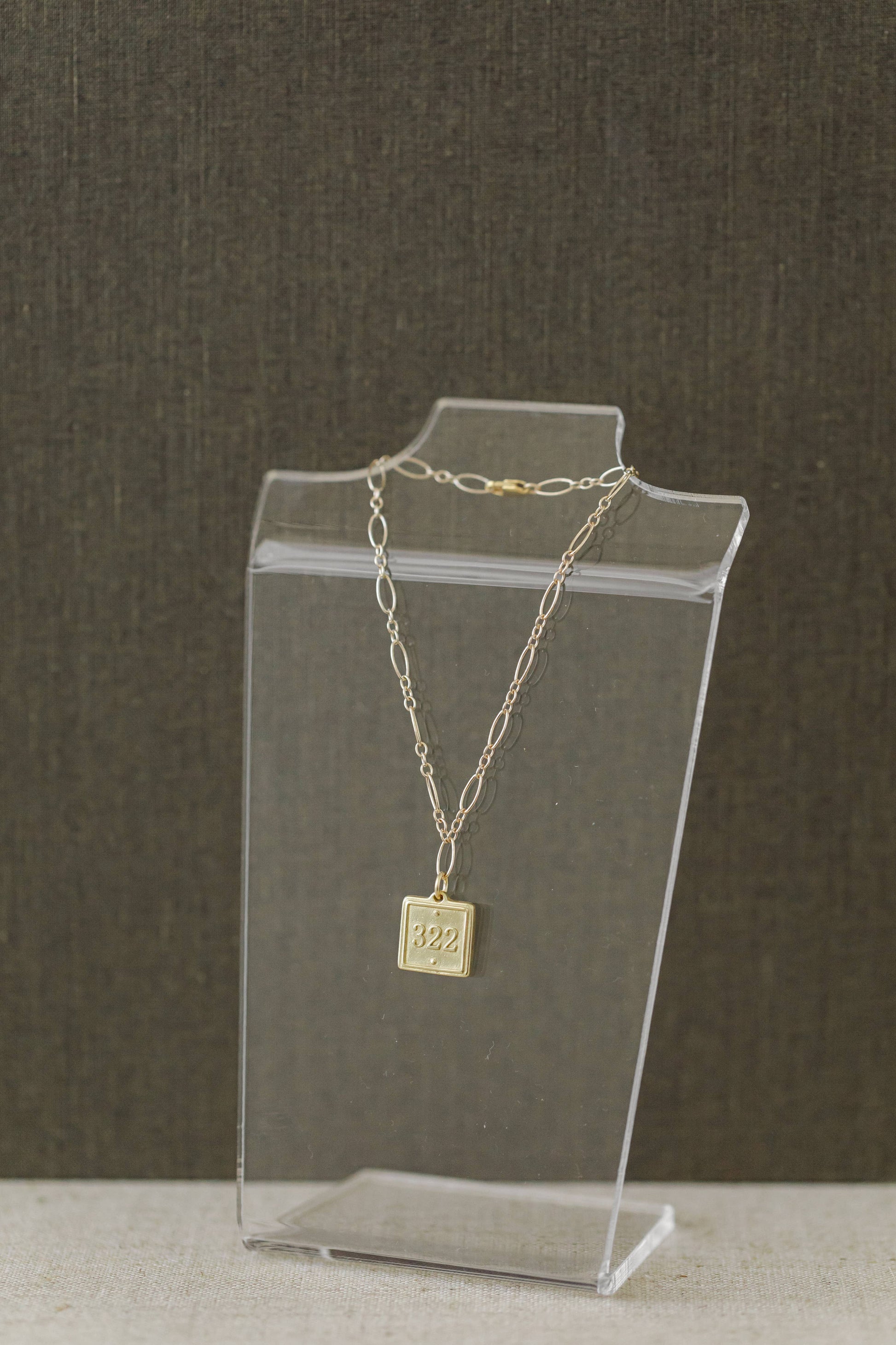 A delicate gold chain necklace with a square pendant engraved with '82$', displayed on a clear acrylic stand against a dark textured background.