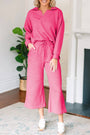 Woman wearing a pink outfit standing in a room with a plant and fireplace.