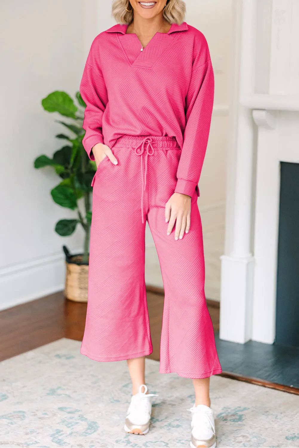 Woman wearing a pink outfit standing in a room with a plant and fireplace.