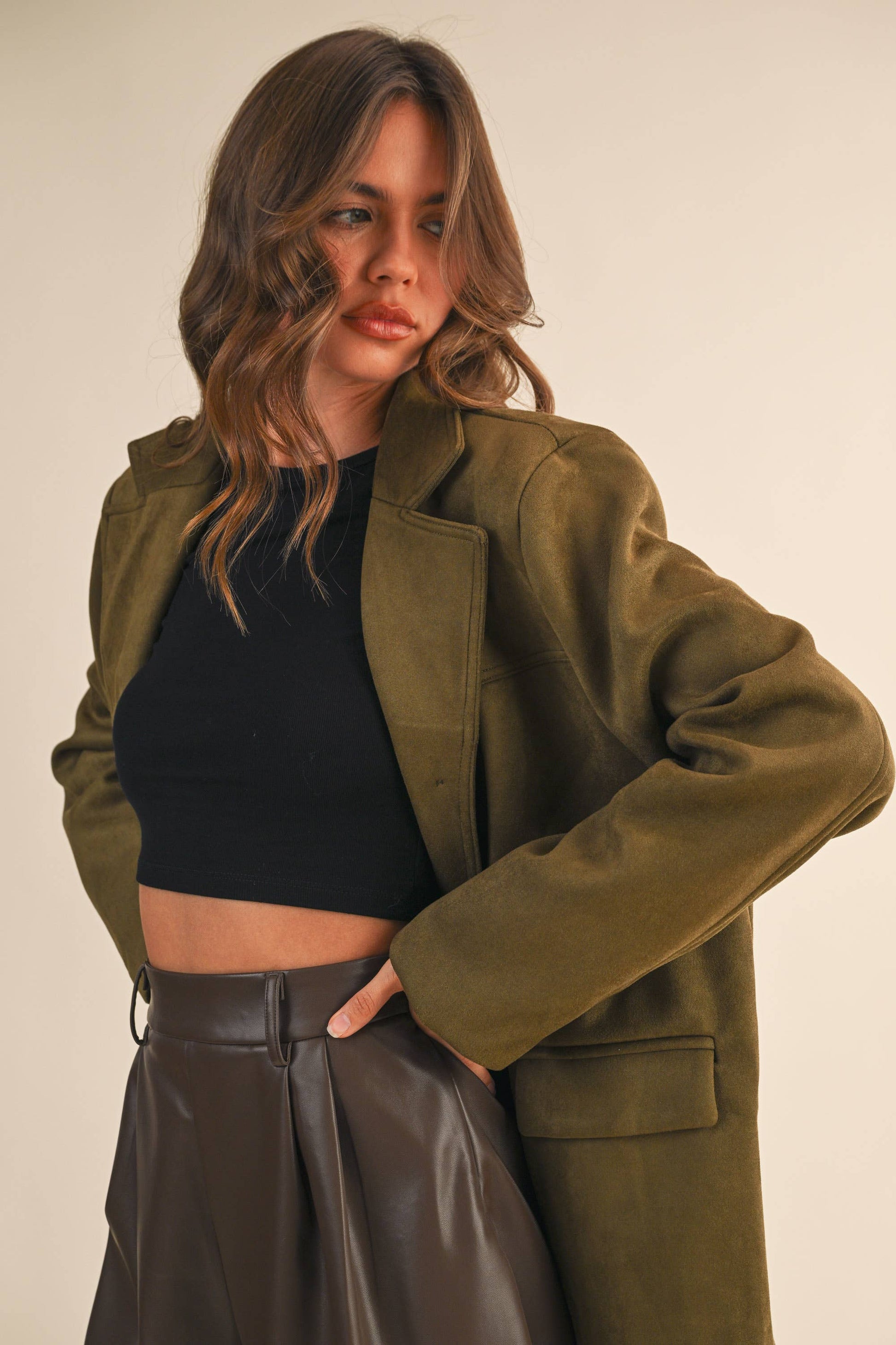 Woman wearing a green suede jacket over a black top and brown shorts on a plain background