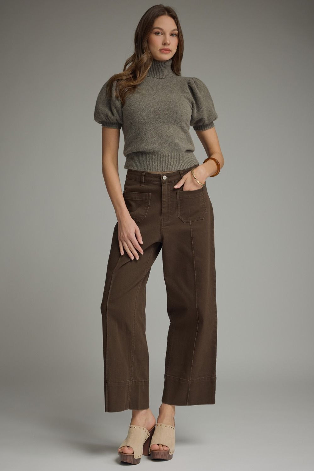 Woman wearing a gray turtleneck and brown pants against a plain background