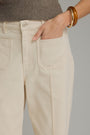 Close-up of a person wearing light beige straight-leg jeans with a ribbed taupe sweater, a gold bangle, and a hand tucked into the front pocket, against a solid gray background.