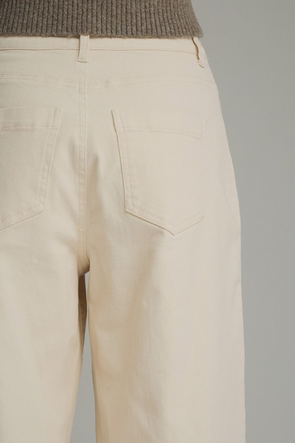 Close-up back view of a person wearing light beige wide-leg trousers with a ribbed taupe knit top visible at the waistband, against a plain light gray studio background.