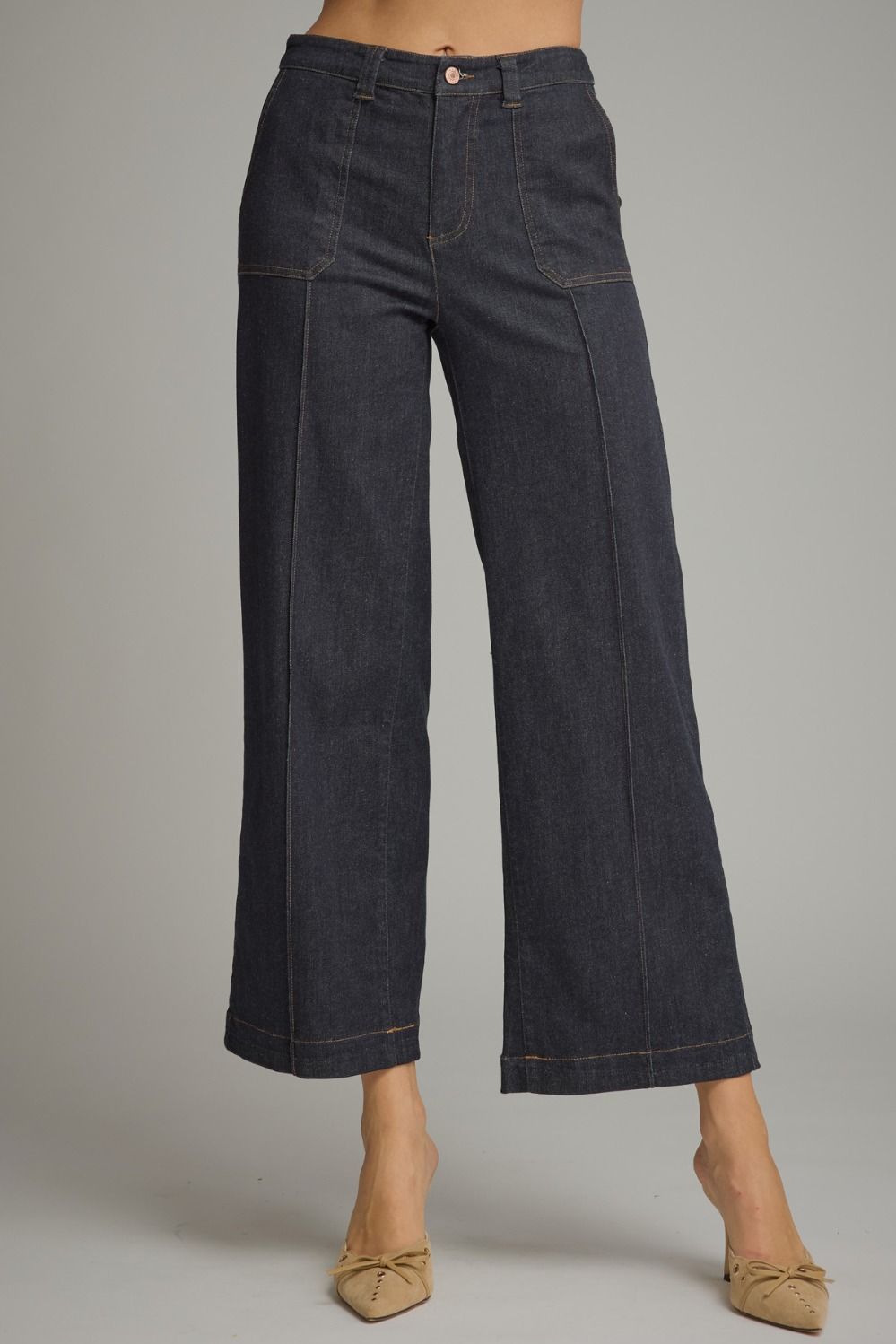 A model wearing dark indigo wide-leg cropped denim pants with beige pointed-toe flats, shown from the waist down against a neutral gray studio background.