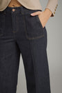 Close-up of a person wearing dark indigo denim jeans with a beige blazer, showing the side pocket, front seam, waistband, and hand in the pocket against a plain gray background.